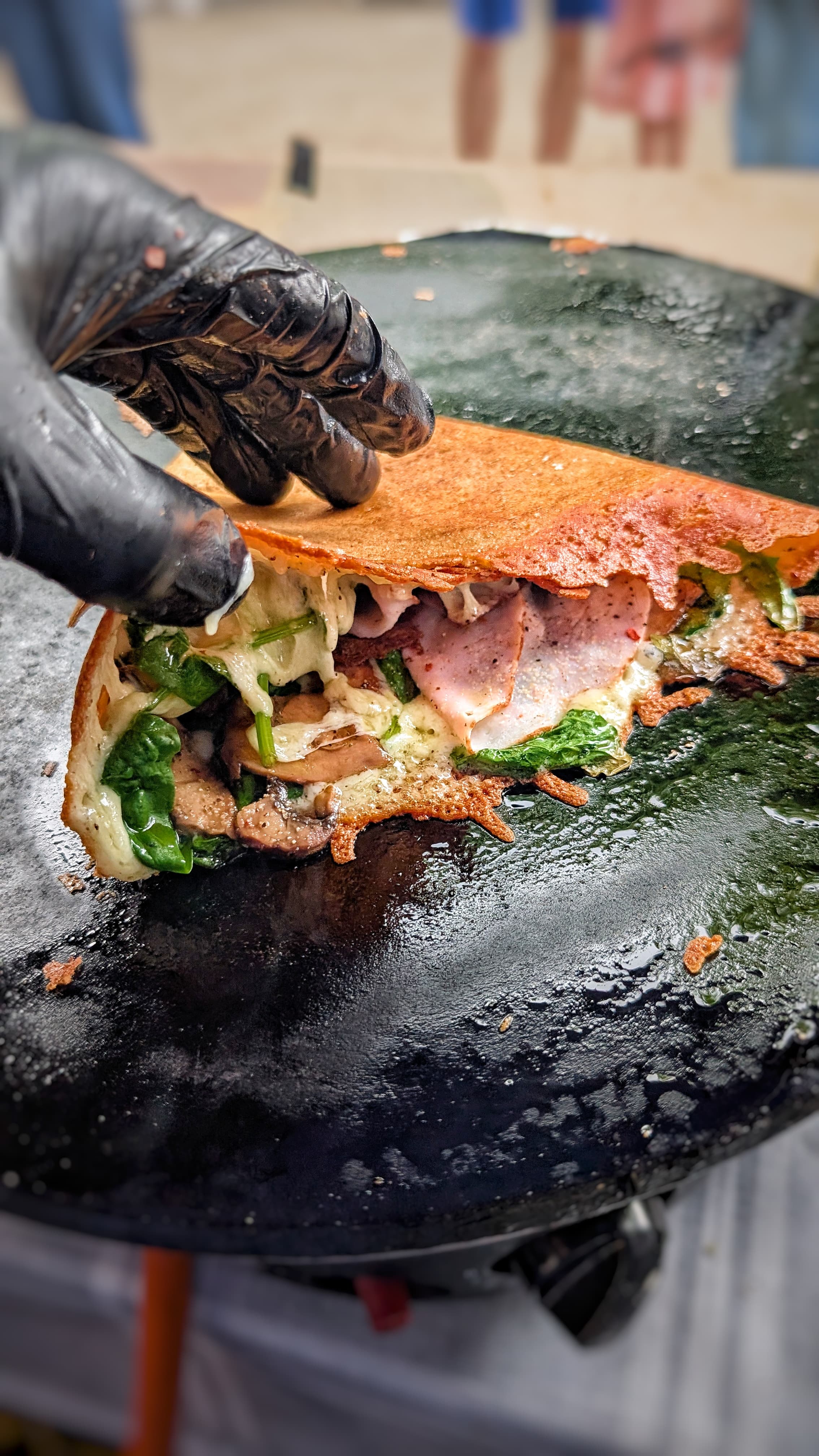 A gloved hand folding a buckwheat crêpe filled with smoked ham and spinach on the stone billig